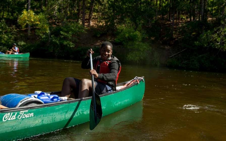 A person wearing a life jacket paddles a canoe on calm water near a tree-lined shore.